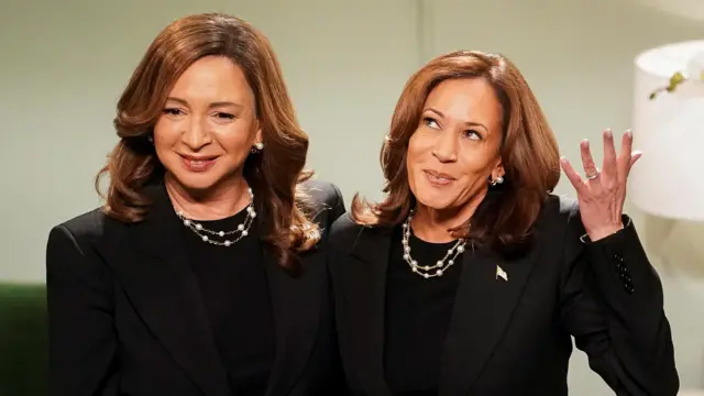 Comedian Maya Rudolph smiles next to Kamala Harris who makes a whimsical gesture - they're dressed identically