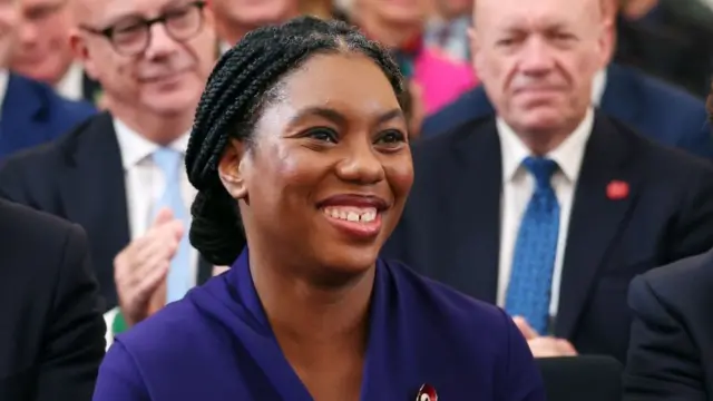 Close up shot of Kemi Badenoch reacting after she's proclaimed leader. She's wearing a blue dress with the top of a enamel poppy bin visible below her left shoulder. Behind her are two men in suits, slightly blurry