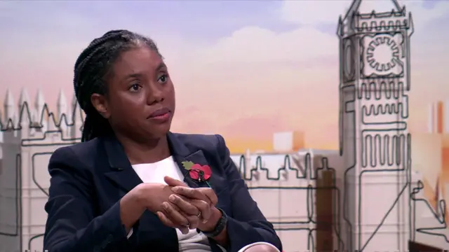 Kemi Badenoch in black jacket and white shirt sitting down on chair. In background, is a carton version of the House of Parliament with the Big Ben to the right of the snap