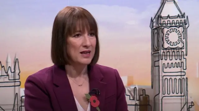 Rachel Reeves in plum suit, white blouse and poppy pin on her left lapel sitting down. Behind her is a cardboard model of the House of Parliament with Big Ben