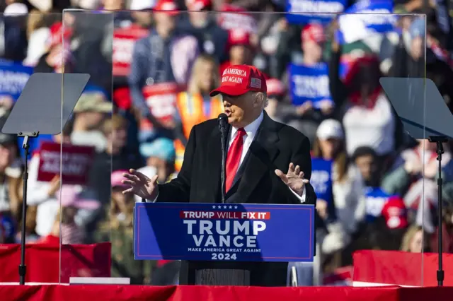 Donald Trump, in a MAGA hat, stands behind safety glass at a rally