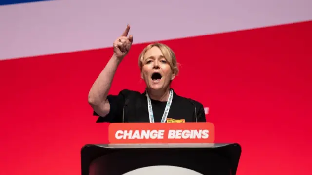 General Secretary of the Unite union, Sharon Graham speaks during a debate. She's speaking behind a lectern with a sign reading "change begins" on red cardboard. Her right hand is raised and she has her pointer finger up as she speaks. She's in a black dress