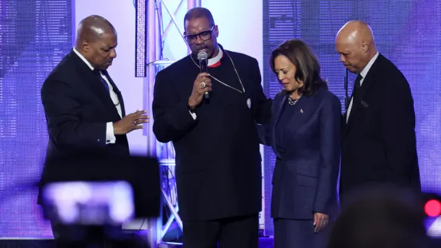 A minister speaks into a microphone, as he locks arms with Kamala Harris. Two men stand either side, also in prayer