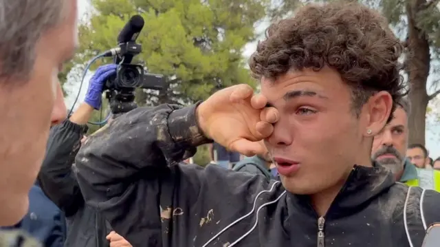 Sixteen-year-old Pau rubs his right eye with his right hand as he cries. His black jacket and hands have been scattered in mud