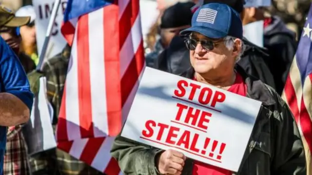 A man, wearing sunglasses, a red tshirt, grey jacket and a blue cap with an American flag, holds a Stop The Steal sign