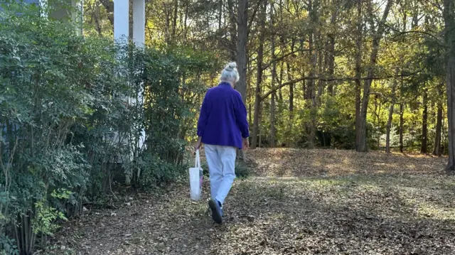 Woman walks through leave-filled grass towards an area of trees in front of her. She is wearing a bright blue cardigan and jeans, and carrying a white bag in her left hand