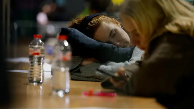 Two women sit behind a desk - one is on her phone and the other is asleep.