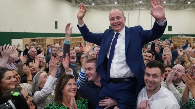 Micheál Martin, wearing a suit, smiling and raising his hand in the air