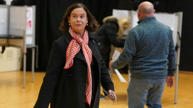 Mary Lou McDonald, wearing a red and white scarf and a black coat, walking through a voting centre. There is a man walking behind her.