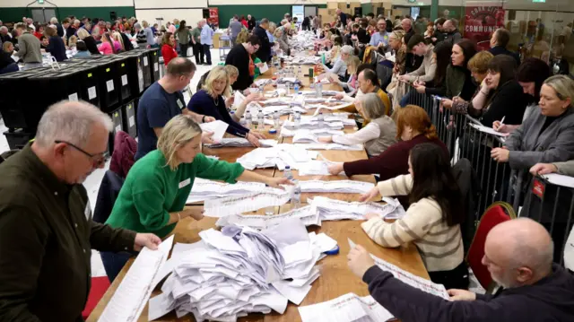 A long table filled with ballot papers. There are people lined up on either side of the table