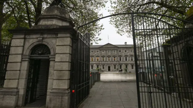 A black gate in front of a driveway going up to a large stone building that is three storeys high.