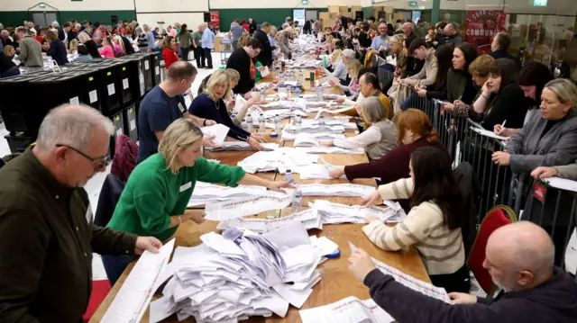 People standing at either side of a long table full of ballot papers.