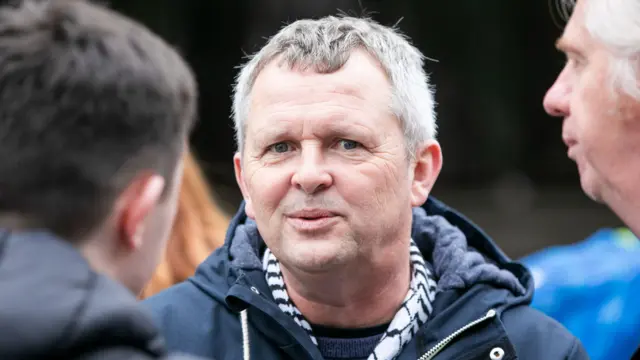 A man with grey hair looking at the camera. He is wearing a navy coat.