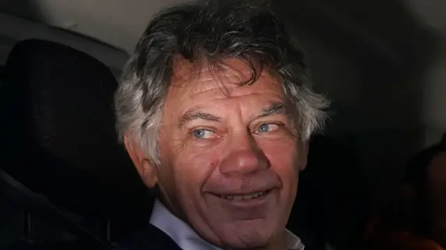 A man, looking to the left of the frame and smiling. He has grey hair.