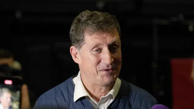 Eamon Ryan, a man with short dark brown hair, pictured from the shoulders up. He is wearing a white shirt and navy top. The background of the image is dark.