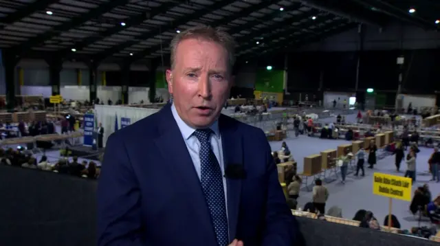 A man, wearing a navy suit, standing in an election count centre.