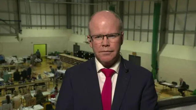 Peadar Tóibín, wearing a navy suit, white shirt and red tie. He is also wearing glasses. He is standing in an election count centre, visible in the background.