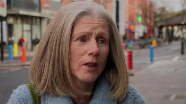 Debra Nolan speaks to a reporter, who is behind the camera. She has short blonde hair cut into a bob. She has blue eyes and is wearing a fluffy light blue coat. She is stood on the pavement on a street with brick buildings in the background.