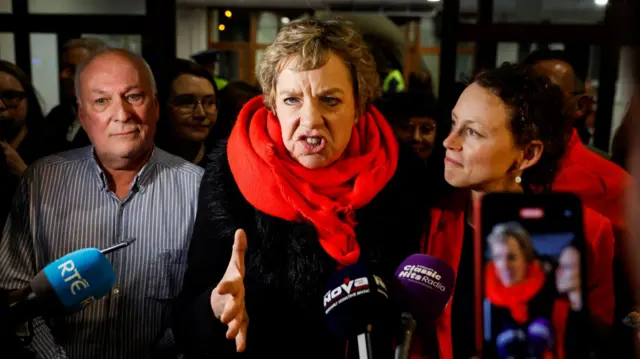 A woman, with short blonde hair, wearing a black jacket and red scarf. She is standing beside a man and a woman. There are media microphones in front of her.