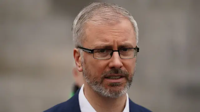 Roderic O'Gorman, pictured from the shoulders up. He has a tight grey beard and grey hair. He is wearing glasses. The background of the image is blurred.