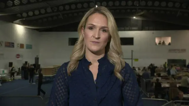 A woman with blonde hair, wearing a navy top. She is standing in an election count centre, visible in the background.