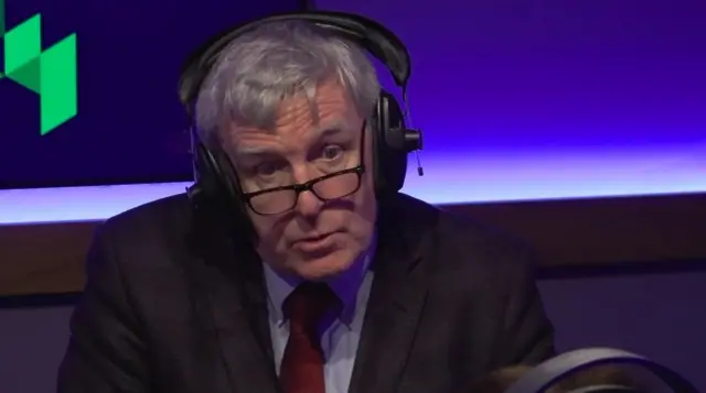 A man with grey hair, wearing a suit, sitting in a radio studio. He is wearing glasses and headphones.