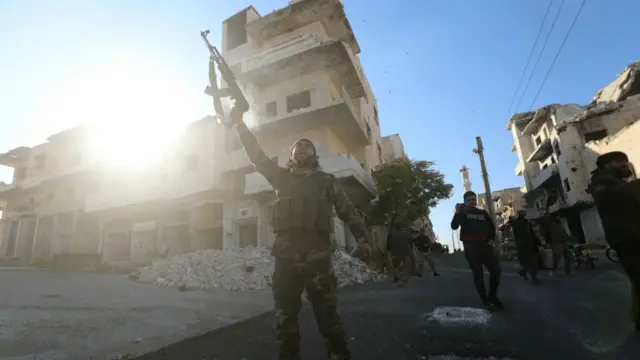 An anti-government fighter raises his gun against the backdrop of damaged buildings