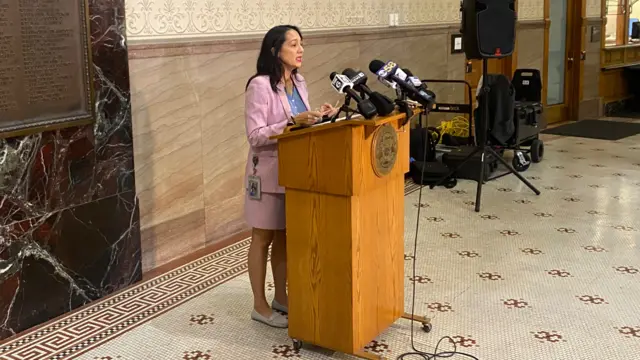 Paulina Guiterrez standing in front of a lectern. She is wearing a pink suit with a blue shirt, and she is speaking into press microphones