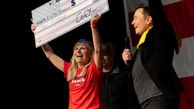 A woman holds up a large cardboard cheque for one million dollars, standing next to Elon Musk on stage