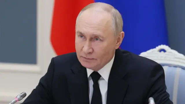 Vladimir Putin, sitting in front of a Russia flag, looks off to his right