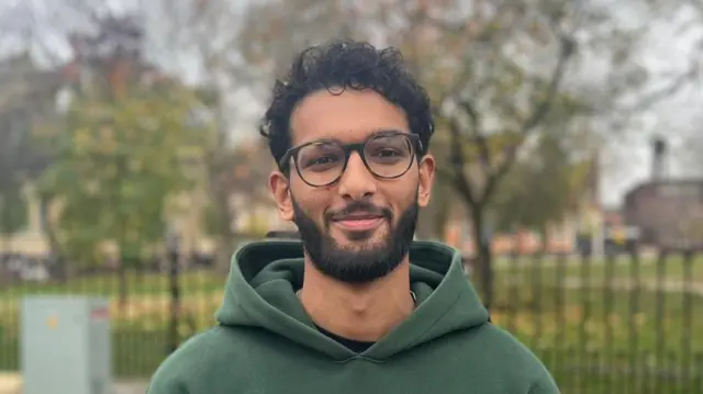 Zay, Manchester Met University student, stands looking to camera with a dark green hoodie on. He is wearing glasses and is smiling. He has short, curly black hair and black facial hair