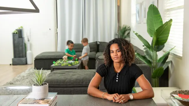 Allison McCullough sits in a living room, with two children playing games in the background