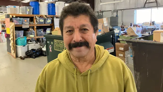 Pedro Fernandez in a warehouse. He is standing in front of stacks of boxes of donations and he is wearing a yellow hoodie.