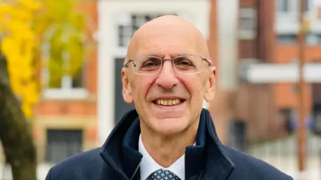 Professor Malcolm Press stands looking to camera and smiling. He is wearing a navy blue coat, white shirt and blue shaded patterned tie