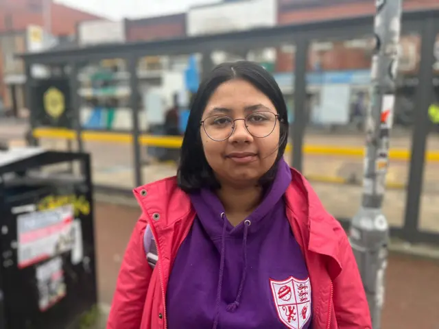 Laaibah, Manchester Met student, is seen looking at the camera with a neutral expression. They are wearing a pink coat and purple hoodie with a crest on it. They have black hair and glasses.