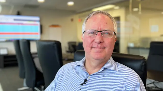 Joe Lenski, dressed in a blue and white shirt, smiles into the camera in an office, with a spreadsheet on a TV in the background