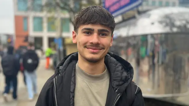 Shay, student at Manchester Met University stands looking to the camera and smiling. He is holding a coffee cup in his right hand and is wearing a black jacket and dark beige t-shirt. He has medium length brown hair and a moustache