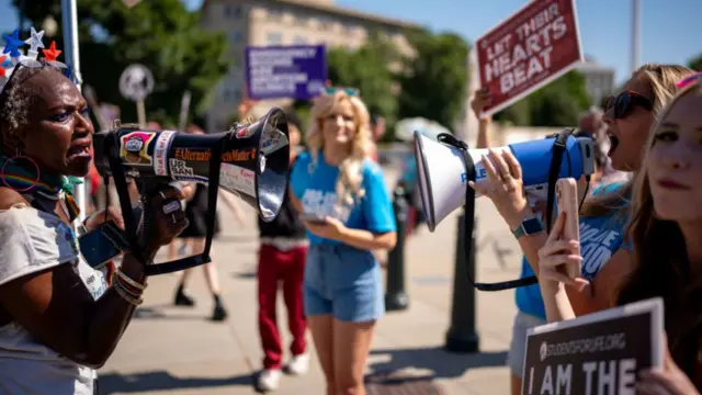 Two people with megaphones facing each other and shouting at each other. People with signs for and against abortion stand around them