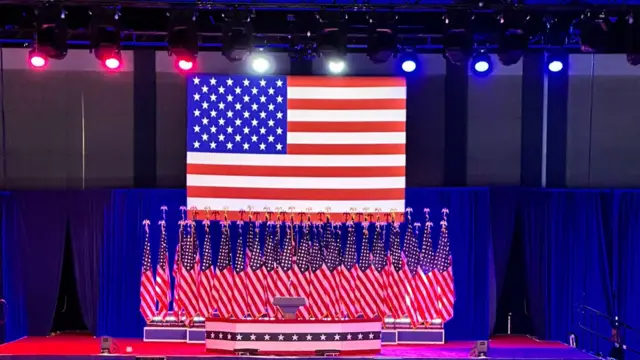 A stage with red, blue and white spotlights, a podium, a huge American flag and then several more American flags