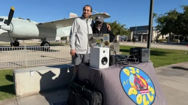 Two men hug behind their DJ booth which says DJs at the polls on the front of it. An aeroplane is parked behind them