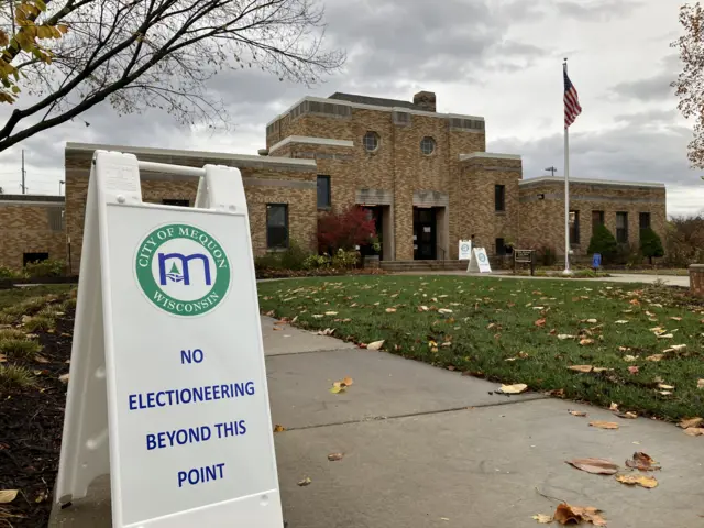 A picture of the Mequon City Hall in the background. In front of it is an American flag. In the foreground is a sign that reads 'no electioneering beyond this point'