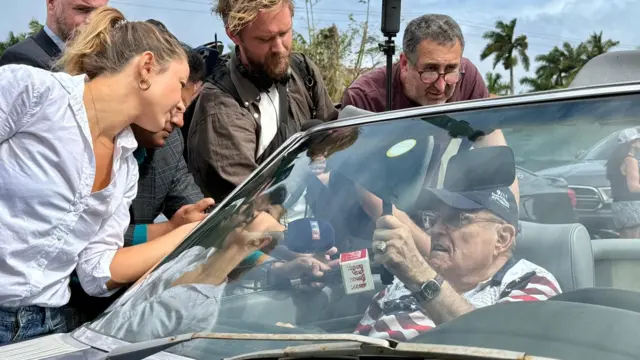 Rudy Giuliani in a convertible surrounded by reporters putting microphones in his face