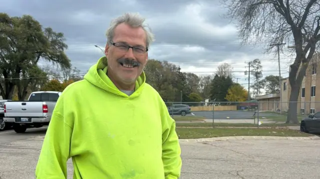 A man stands in a parking lot in a green sweater