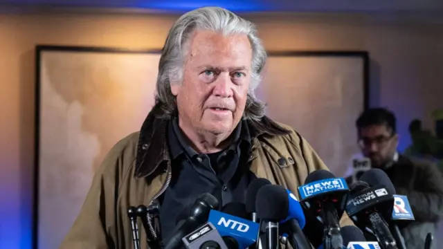 Steve Bannon, wearing a brown jacket and black shirt, stands at a podium containing several microphones as he takes questions at a press conference