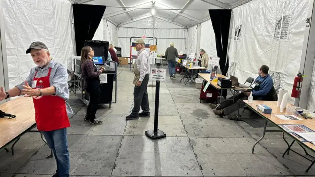 A picture of a tent with some tables in it and people stood around