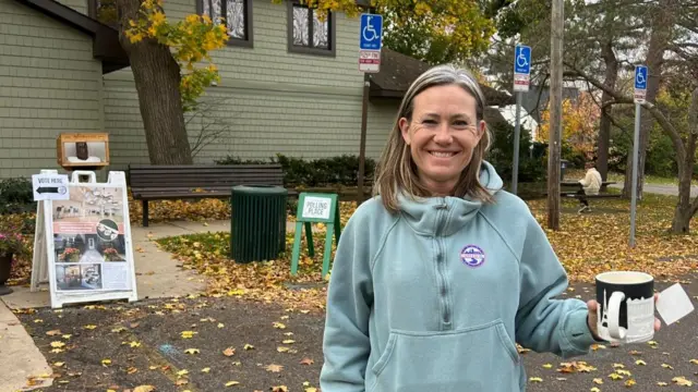 A woman smiling holding a mug in front of a polling station