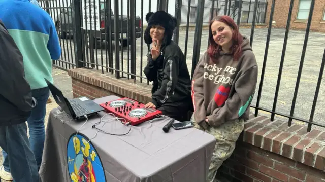 Two people stand behind a makeshift DJ booth outside - a table with some decks on it - while others queue nearby