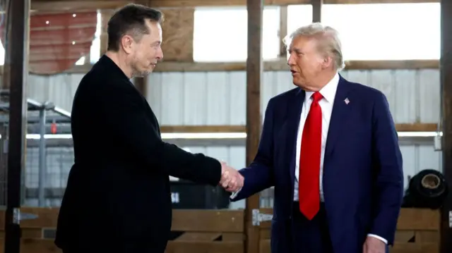 Elon Musk shakes hands with Donald Trump, who is wearing a blue suit with a red tie