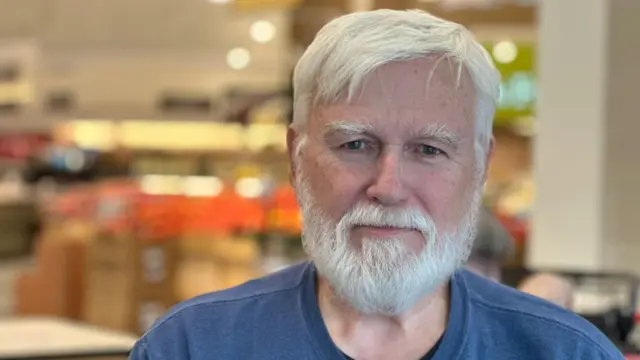 Headshot of Arthur smiling at camera, he wears a blue tshirt and has grey beard and facial hair