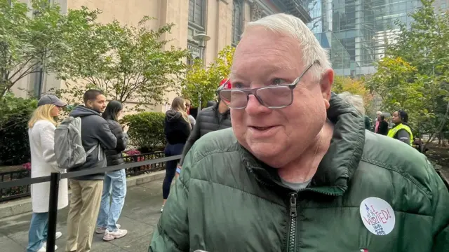 Headshot of Francis talking to the camera. There's a line behind him. He is wearing a green puffer jacket with a sticker on it that says 'I voted'